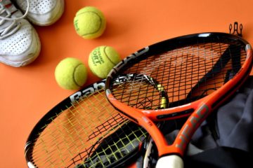 Some tennis rackets, a bag, balls, and shoes on an orange floor intended to show off some of the tennis accessories being reviewed as part of an introductory post on a tennis review website.